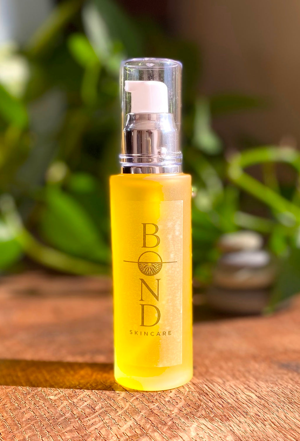 Bond Recovery Body Oil in an amber frosted glass pump bottle on a wooden surface, with green plants and stacked stones behind