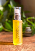 Bond Recovery Body Oil in an amber frosted glass pump bottle on a wooden surface, with green plants and stacked stones behind