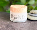 Bond Whipped Tallow Cream in a frosted glass jar with a wooden lid, beside stacked stones and greenery