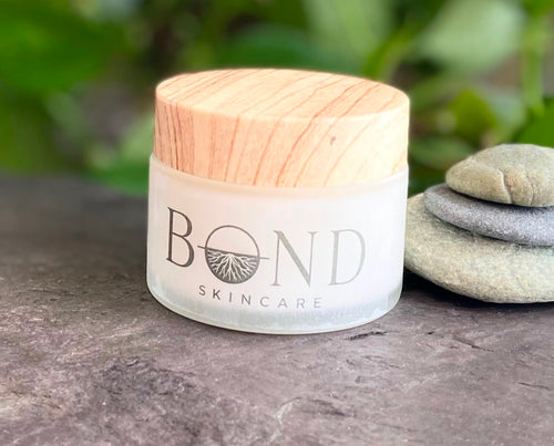 Bond Whipped Tallow Cream in a frosted glass jar with a wooden lid, beside stacked stones and greenery