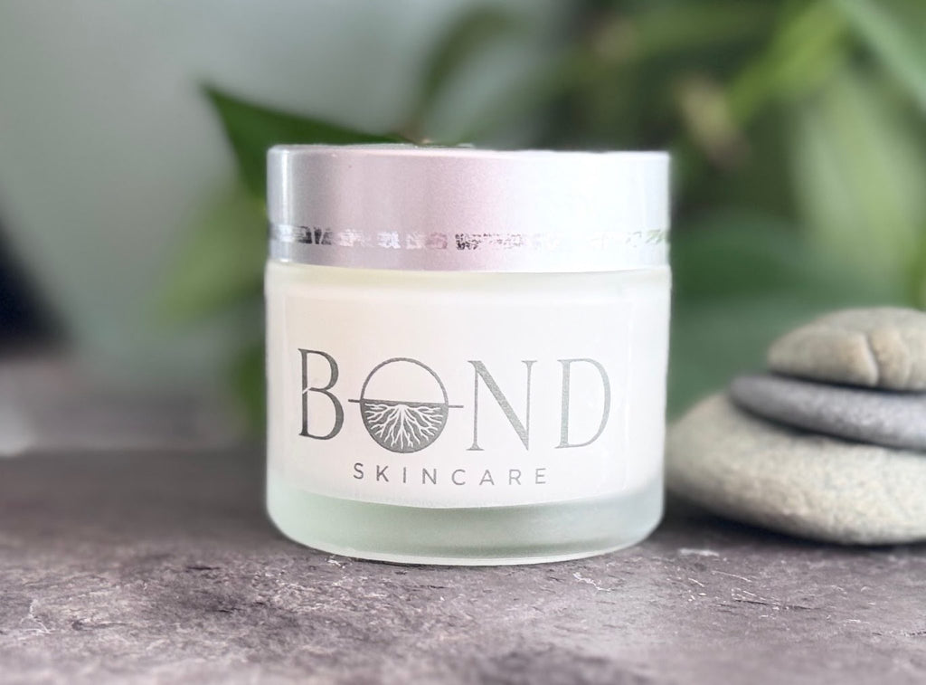 Close-up of the Bond Nectar Serum frosted glass jar with a silver lid beside stacked stones