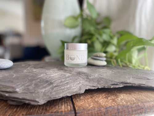 Bond Nectar Serum in a frosted glass jar with a silver lid on a slate stone beside stacked pebbles and green plants