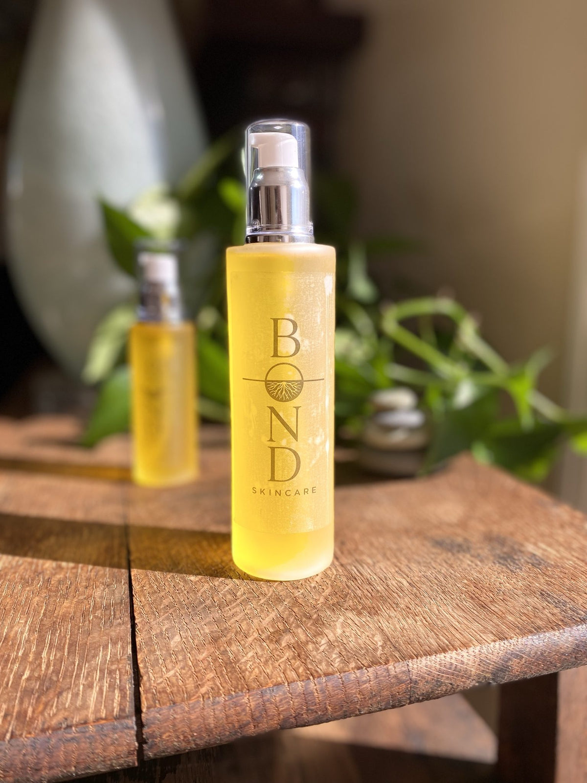 Bond Recovery Oil large pump bottle in front of the smaller 2 oz bottle on a wooden table with green plants