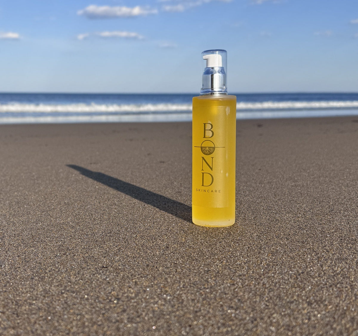 Bond Recovery Oil in a large amber pump bottle standing on a sandy beach with the ocean and blue sky behind