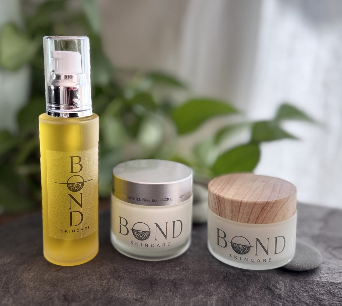 Bond Recovery Oil bottle, Nectar Serum jar, and Whipped Tallow Cream jar together on slate in front of green plants
