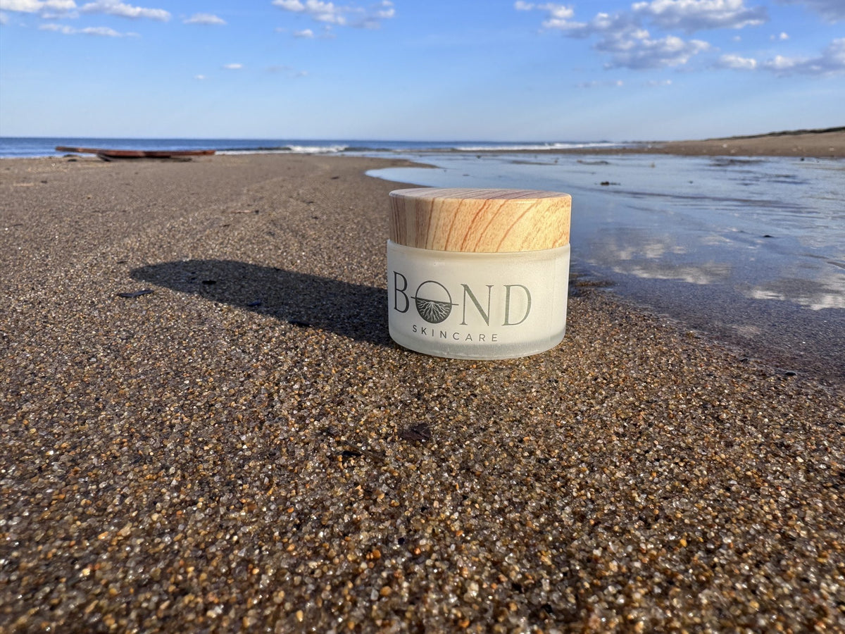 Bond Whipped Tallow Cream jar with a wooden lid resting on wet sand at the edge of a tide pool under a blue sky