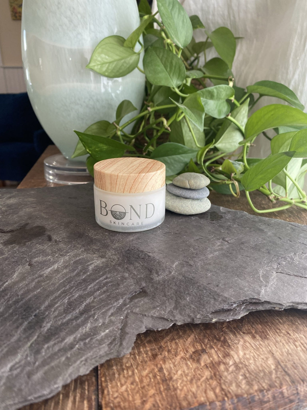 Bond Whipped Tallow Cream jar with a wooden lid on a slate stone, with green plants and stacked pebbles behind